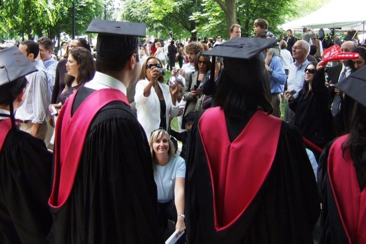Graduates in black robes and red hoods gather outdoors at a graduation ceremony.