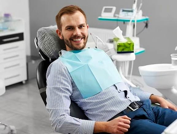 Smiling male patient wearing blue bib relaxing in dental chair at modern dentistry office.