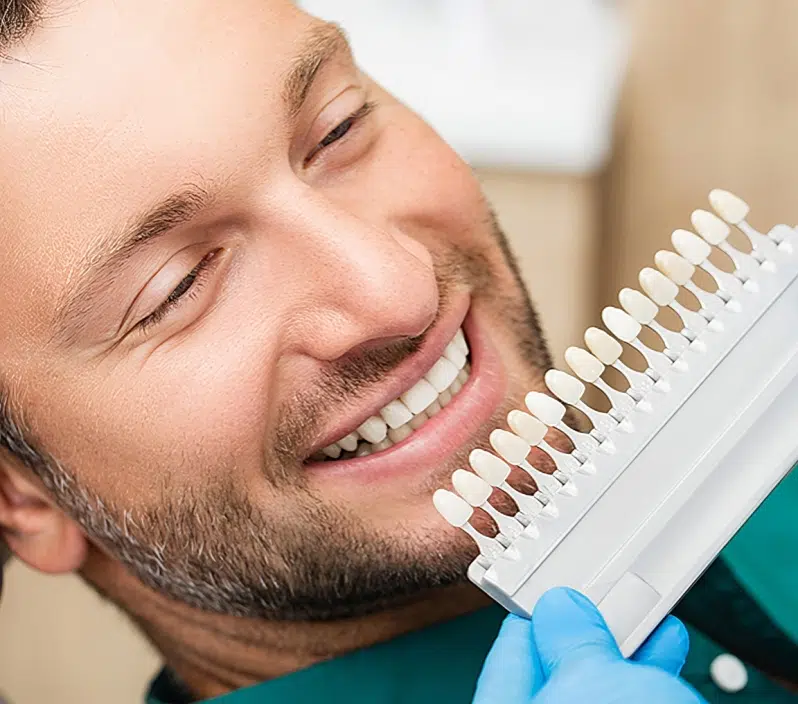 Man smiling while holding dental shade guide during teeth whitening consultation at dentistry office.