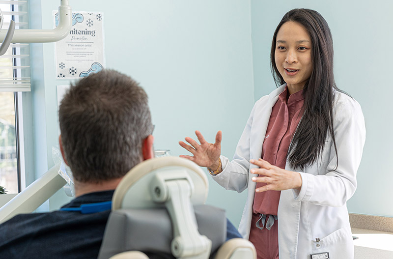 Female dentist in white coat gestures while consulting with male patient in dental office.