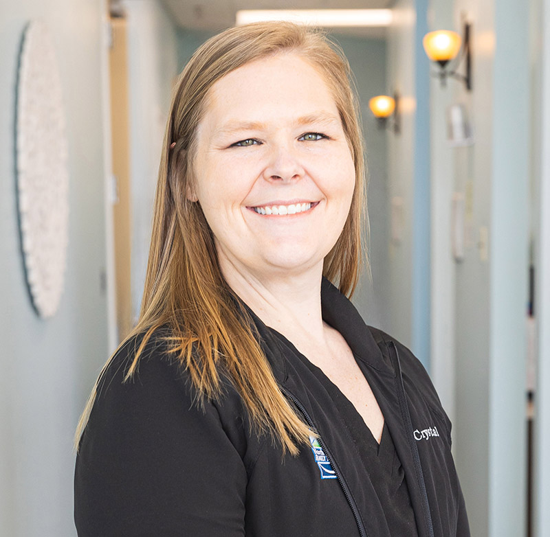 Professional woman smiling in dental office hallway wearing black scrubs with name badge.