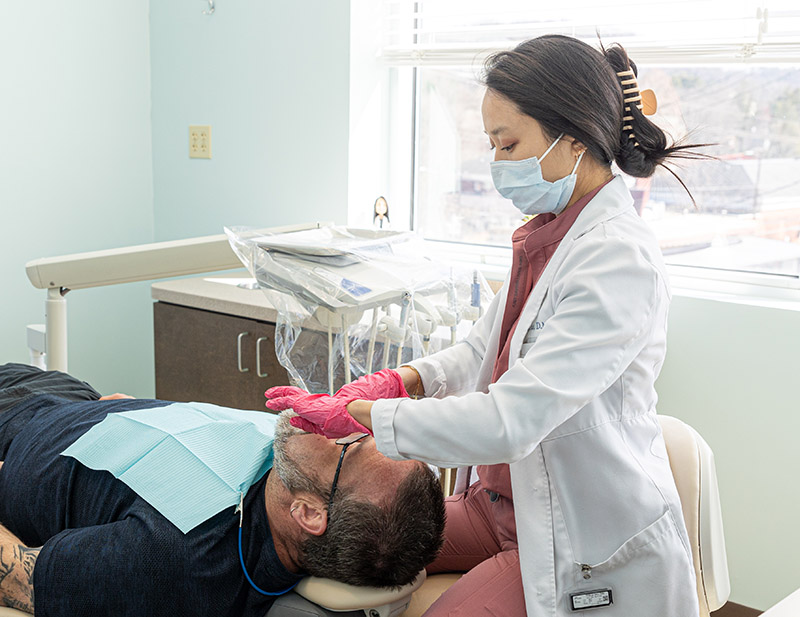 Dentist in white coat and mask examines patient's teeth in modern dental clinic operatory.