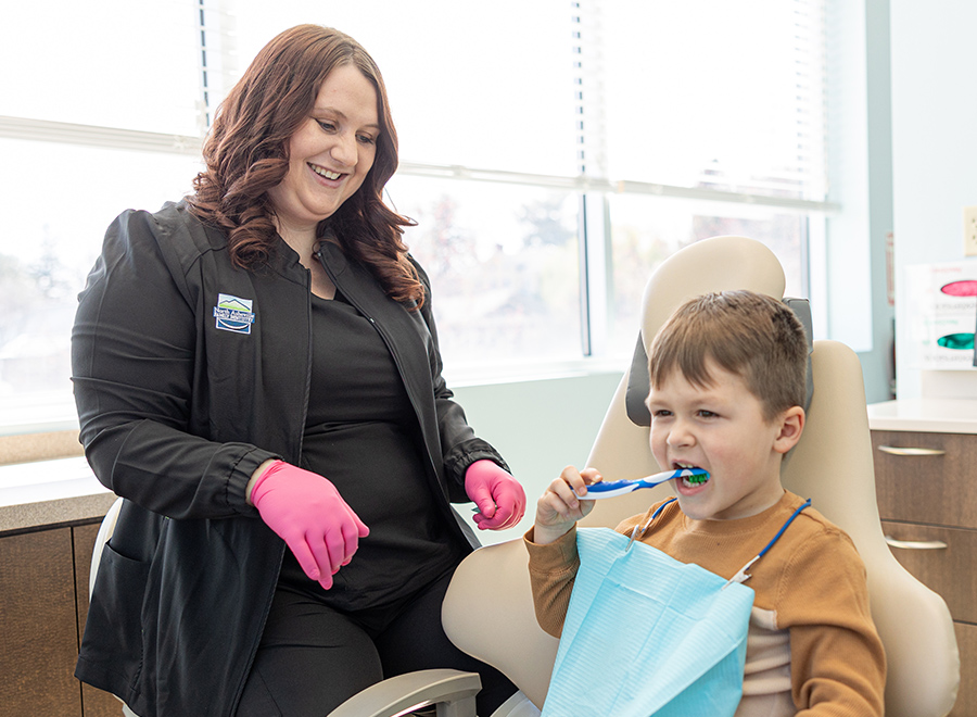 Female dental professional with pink gloves helping young boy brush teeth in dental office.
