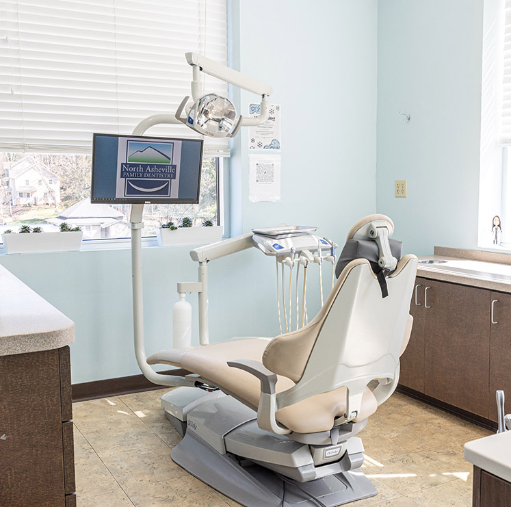 Modern dental treatment room with empty patient chair, overhead light, monitor, and equipment.
